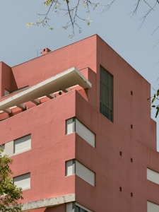 A modern building with a minimalist architectural design characterized by a bold pink exterior and a series of rectangular windows. The structure includes a cantilevered white balcony with clean lines. In the foreground, green foliage partially obscures the lower part of the building, while thin branches with sparse leaves frame the image at the top.