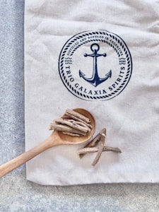 Canvas cloth with a navy blue circular logo featuring an anchor and the words 'TRIO GALAXIA SPIRITS ESTD 2022'. A wooden spoon holding dried sticks is placed on the cloth.
