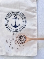 A light-colored fabric bag with a navy-blue print featuring an anchor and text. A wooden spoon rests on the bag, filled with small round seeds, with some scattered around the spoon.