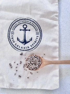 A light-colored fabric bag with a navy-blue print featuring an anchor and text. A wooden spoon rests on the bag, filled with small round seeds, with some scattered around the spoon.