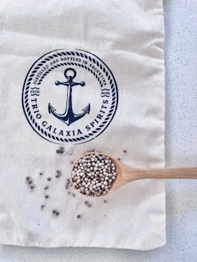 A light-colored fabric bag with a navy-blue print featuring an anchor and text. A wooden spoon rests on the bag, filled with small round seeds, with some scattered around the spoon.