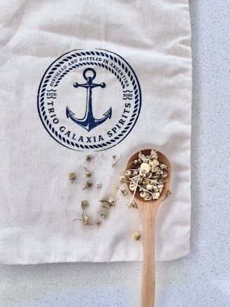 A wooden spoon filled with dried chamomile flowers rests on a beige fabric with a blue anchor logo and text 'Trio Galaxia Spirits'. The fabric sits on a speckled, light grey surface.