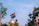 A hand wearing a rainbow wristband and fishnet glove holds up a rainbow flag. The background features trees, a building, and a construction crane under a clear blue sky.