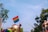 A hand wearing a rainbow wristband and fishnet glove holds up a rainbow flag. The background features trees, a building, and a construction crane under a clear blue sky.