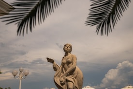 A weathered stone statue of a mythological figure stands prominently against a partly cloudy sky. The figure appears to be holding a draped cloth, with an arm extended outward. Palm fronds frame the upper corners of the image, providing a tropical atmosphere. A decorative street lamp with spherical lights can be seen to the left of the statue.