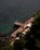 A wooden deck stretches out over aqua blue seawater, lined with sun loungers and orange umbrellas. The deck is bordered by lush green vegetation and dark rocky formations on one side, while the calm sea extends on the other. A few people can be seen relaxing on the loungers.