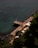 A wooden deck stretches out over aqua blue seawater, lined with sun loungers and orange umbrellas. The deck is bordered by lush green vegetation and dark rocky formations on one side, while the calm sea extends on the other. A few people can be seen relaxing on the loungers.