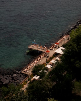 A wooden deck stretches out over aqua blue seawater, lined with sun loungers and orange umbrellas. The deck is bordered by lush green vegetation and dark rocky formations on one side, while the calm sea extends on the other. A few people can be seen relaxing on the loungers.