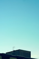 A minimalist image featuring the top of a building with a visible metal ladder against a clear blue sky. The structure is angular and industrial, with clean lines and a modern appearance.