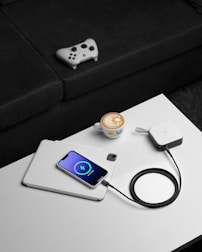 A white smartphone is charging on top of a tablet on a white table. The phone displays a charging symbol on the screen. Next to it, there's a charging device connected with a black cable. On the table, a cup of coffee with latte art is placed close to the devices. In the background, a video game controller rests on a black sofa.