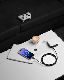 A white smartphone is charging on top of a tablet on a white table. The phone displays a charging symbol on the screen. Next to it, there's a charging device connected with a black cable. On the table, a cup of coffee with latte art is placed close to the devices. In the background, a video game controller rests on a black sofa.