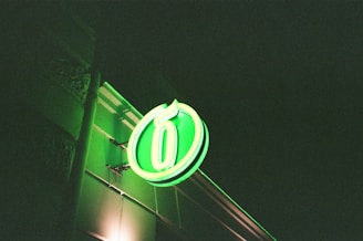 A vibrant green neon sign is mounted on the side of a building, displaying the number six within an oval shape. The surrounding area is dark, highlighting the glow of the neon light and casting a green hue on the adjacent wall.