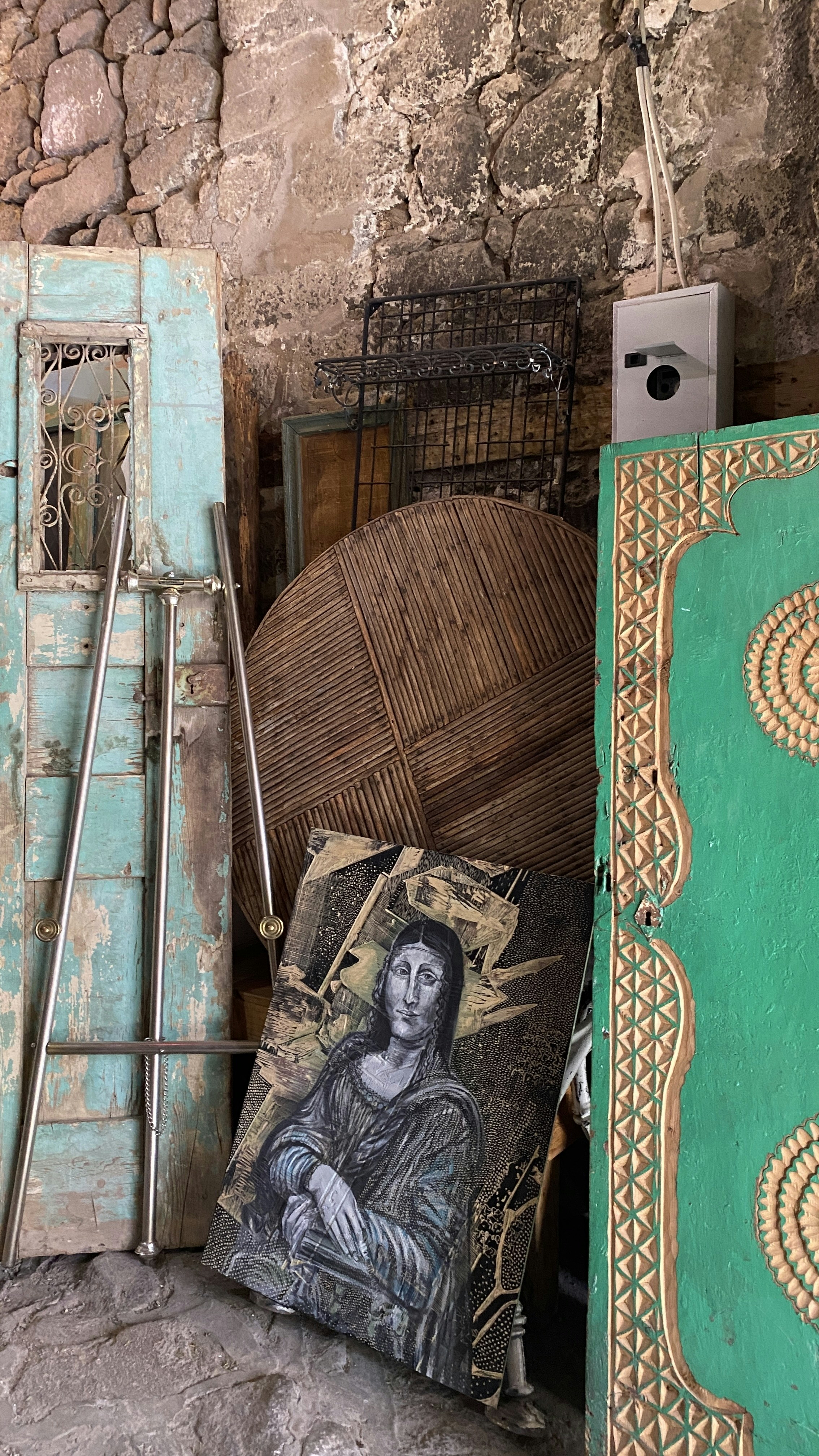 A collection of rustic objects is leaning against a stone wall. There are two vintage doors, one in distressed turquoise and the other in bright green with intricate designs. A large, round rattan piece and other metal elements are stacked among them. A canvas featuring a stylized portrait that resembles the Mona Lisa is prominently placed at the forefront.
