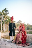A couple dressed in traditional wedding attire smiling at each other, standing outdoors near a body of water. Tall palm trees and a clear sky fill the background, creating a serene and picturesque setting.