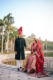 A couple dressed in traditional wedding attire smiling at each other, standing outdoors near a body of water. Tall palm trees and a clear sky fill the background, creating a serene and picturesque setting.