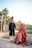 A couple dressed in traditional wedding attire smiling at each other, standing outdoors near a body of water. Tall palm trees and a clear sky fill the background, creating a serene and picturesque setting.