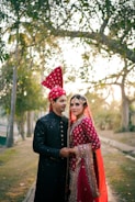 A couple dressed in traditional wedding attire stands closely together. The woman wears a bright red embroidered dress with intricate patterns and a matching headpiece, while the man is dressed in a black sherwani with subtle embellishments and a red turban. They are surrounded by trees and soft sunlight filtering through, creating a serene and romantic atmosphere.