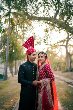 A couple dressed in traditional wedding attire stands closely together. The woman wears a bright red embroidered dress with intricate patterns and a matching headpiece, while the man is dressed in a black sherwani with subtle embellishments and a red turban. They are surrounded by trees and soft sunlight filtering through, creating a serene and romantic atmosphere.