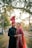 A couple dressed in traditional wedding attire stands closely together. The woman wears a bright red embroidered dress with intricate patterns and a matching headpiece, while the man is dressed in a black sherwani with subtle embellishments and a red turban. They are surrounded by trees and soft sunlight filtering through, creating a serene and romantic atmosphere.