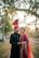 A couple dressed in traditional wedding attire stands closely together. The woman wears a bright red embroidered dress with intricate patterns and a matching headpiece, while the man is dressed in a black sherwani with subtle embellishments and a red turban. They are surrounded by trees and soft sunlight filtering through, creating a serene and romantic atmosphere.