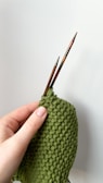A hand is holding two wooden knitting needles and working on a piece of green knitted fabric with a bumpy texture.