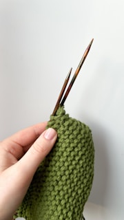 A hand is holding two wooden knitting needles and working on a piece of green knitted fabric with a bumpy texture.