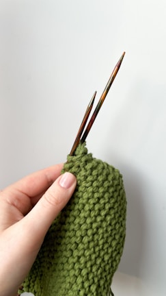 A hand is holding two wooden knitting needles and working on a piece of green knitted fabric with a bumpy texture.