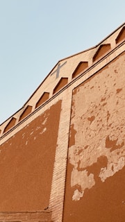 A close-up view of an exterior building wall showcasing architectural features. The wall displays signs of erosion and wear, with visible peeling paint and cracks. There is a vertical brick pattern running along the center. The upper part of the building features a set of small arches under the roofline and a large cross, hinting that it might be a religious building. The background is a clear, light blue sky.