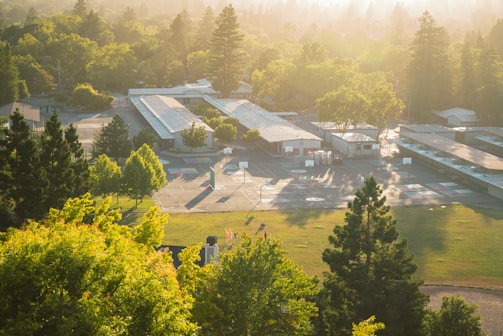 An aerial view of a school campus surrounded by trees and greenery. The campus includes several long, rectangular buildings and a playground area with basketball courts. The scene is bathed in the warm, golden light of sunrise or sunset, casting long shadows across the grounds.