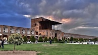 A historic building with intricate architecture and arches under a dramatic sky with patches of sunlight. The foreground features a well-maintained garden with trees and grass, and several people are walking or sitting, enjoying the outdoor space. The scene conveys a blend of cultural heritage and nature.