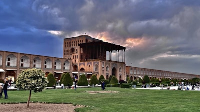 A historic building with intricate architecture and arches under a dramatic sky with patches of sunlight. The foreground features a well-maintained garden with trees and grass, and several people are walking or sitting, enjoying the outdoor space. The scene conveys a blend of cultural heritage and nature.