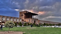 A historic building with intricate architecture and arches under a dramatic sky with patches of sunlight. The foreground features a well-maintained garden with trees and grass, and several people are walking or sitting, enjoying the outdoor space. The scene conveys a blend of cultural heritage and nature.