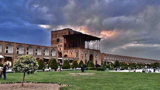 A historic building with intricate architecture and arches under a dramatic sky with patches of sunlight. The foreground features a well-maintained garden with trees and grass, and several people are walking or sitting, enjoying the outdoor space. The scene conveys a blend of cultural heritage and nature.
