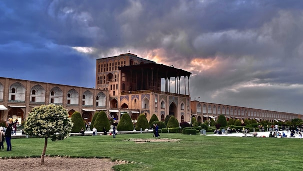 A historic building with intricate architecture and arches under a dramatic sky with patches of sunlight. The foreground features a well-maintained garden with trees and grass, and several people are walking or sitting, enjoying the outdoor space. The scene conveys a blend of cultural heritage and nature.