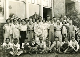 A group of approximately twenty young students and a few adults are gathered for a portrait outside a brick building. The students are dressed in a mix of checkered, floral, and plain clothing typical of a mid-20th-century style. The adults, likely teachers, are positioned behind the group. The image is black and white, suggesting it is from a past era.