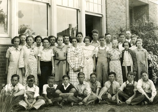 A group of approximately twenty young students and a few adults are gathered for a portrait outside a brick building. The students are dressed in a mix of checkered, floral, and plain clothing typical of a mid-20th-century style. The adults, likely teachers, are positioned behind the group. The image is black and white, suggesting it is from a past era.