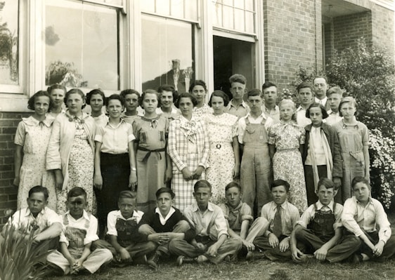 A group of approximately twenty young students and a few adults are gathered for a portrait outside a brick building. The students are dressed in a mix of checkered, floral, and plain clothing typical of a mid-20th-century style. The adults, likely teachers, are positioned behind the group. The image is black and white, suggesting it is from a past era.