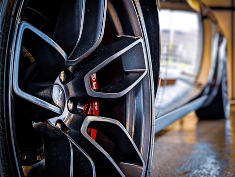 Close-up view of a sleek car wheel with a glossy finish. The focus is on the intricate design of the alloy rim, with visible branding and a vibrant red brake caliper behind it. The reflective surface of the car body can be seen, with blurred reflections of the surrounding environment.