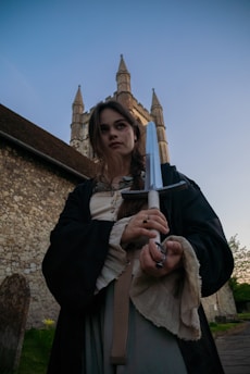 A person dressed in medieval-style clothing holds a sword in a dramatic pose. The background features a historic stone building with towering spires, reminiscent of a castle or cathedral. The sky is clear, giving a serene yet intense atmosphere.