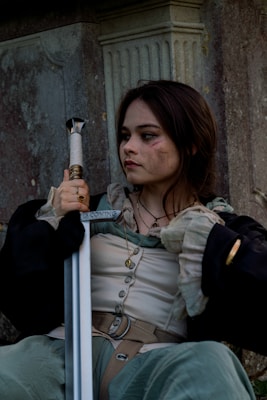 A person with a determined expression, holding a sword, is sitting against a stone column. They are wearing medieval-style clothing and have a few facial scratches, conveying a sense of battle or struggle.