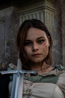 A woman with long hair and a serious expression has subtle facial scars and wears a medieval-style outfit. She holds a sword with the inscription 'Winter is Coming'. The background features a stone pillar, giving a historical or fantasy vibe.