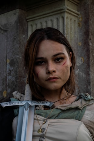 A woman with long hair and a serious expression has subtle facial scars and wears a medieval-style outfit. She holds a sword with the inscription 'Winter is Coming'. The background features a stone pillar, giving a historical or fantasy vibe.