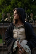A person dressed in historical or medieval-style clothing stands outdoors, holding a dagger. The outfit includes a buttoned blouse, a dark cloak with a hood, and decorative rings. The background features a wrought-iron fence and greenery, suggesting a garden or park setting. The person's expression is contemplative.