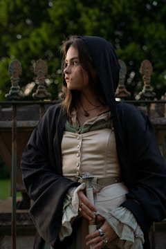 A person dressed in historical or medieval-style clothing stands outdoors, holding a dagger. The outfit includes a buttoned blouse, a dark cloak with a hood, and decorative rings. The background features a wrought-iron fence and greenery, suggesting a garden or park setting. The person's expression is contemplative.