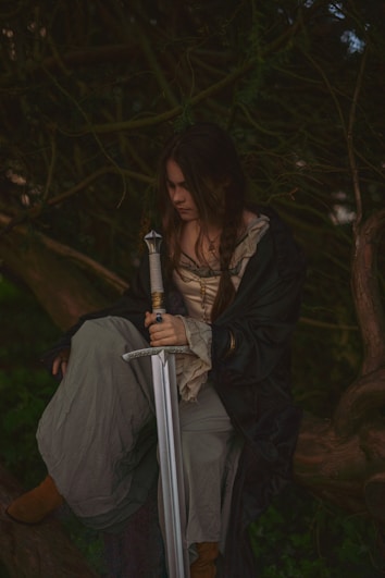 A person with long braided hair sits in a contemplative pose surrounded by dense, dark trees. They are draped in a medieval-style cloak and holding a large, ornate sword with both hands, gazing downward.