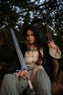 A person with long hair and a serious expression is seated amidst trees, holding a large sword. They wear medieval-style clothing including a dark hooded cloak, a beige corset-like top, and green pants. The setting appears to be outdoors with the backdrop of dense foliage.