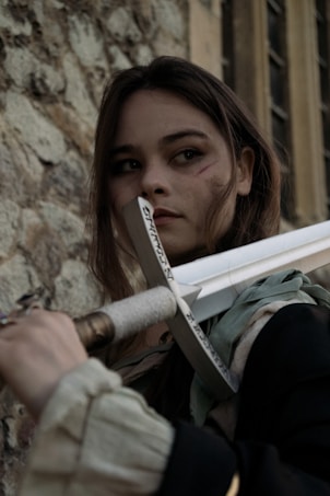 A person with long hair holding a large sword stands against a stone wall, with a focused expression and visible battle marks on the face. The sword has inscriptions on its blade.