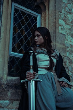 A person dressed in medieval-style clothing holds a sword while leaning against a stone wall beside a window with a metal grid design. The setting appears to be vintage, with the attire consisting of a cloak, a buttoned garment, and loose-fitting pants.