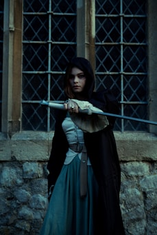 A person with long dark hair is wearing a black hooded cloak, standing in front of a medieval-style stone wall and window with diamond patterns. They are holding a weapon with an intricate handle, projecting an aura of mystery.