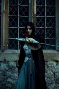 A person with long dark hair is wearing a black hooded cloak, standing in front of a medieval-style stone wall and window with diamond patterns. They are holding a weapon with an intricate handle, projecting an aura of mystery.